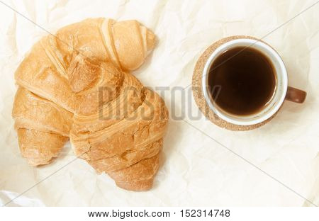 Two baked croissants with a cup of coffee on the white paper's surface.