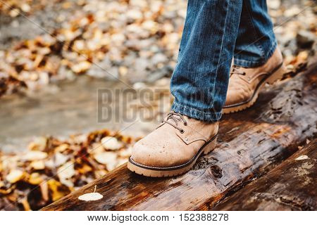 Hiker wearing boots walking across river in autumnal forest. Footwear on man's legs closeup outdoor. Male seasonal shoes.