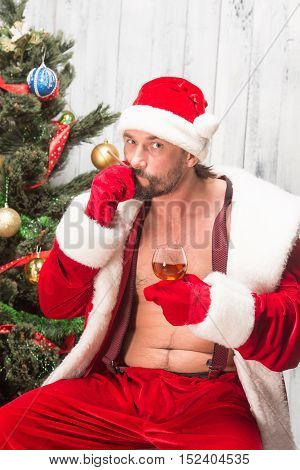 Portrait of happy bad Santa Clause smoking sigar and drinking expensive wine from glass. Handsome Santa Clausa sitting ner New Year Eve or tree.