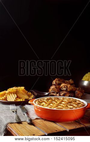 Wooden table served for Thanksgiving dinner. Close view at pumpkin pie. Chips, chicken legs, and mash potatoes on a black background. Vertical shot. Dark photo