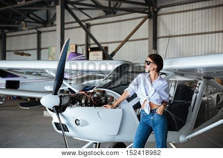 Beautiful young woman in sunglasses posing near plane while standing in angar