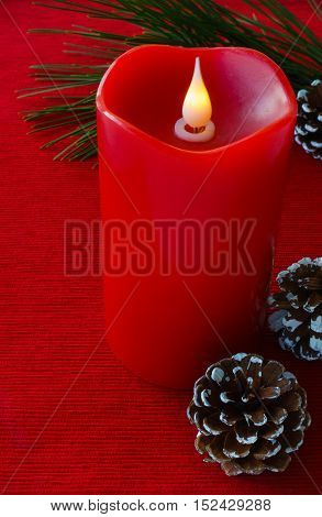 A large red flame less wax candle surrounded by a pine tree twig and pine cones on red fabric background.