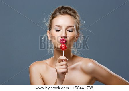 Young beautiful blonde naked girl sending kiss, holding chupa chups over grey background. Copy space.