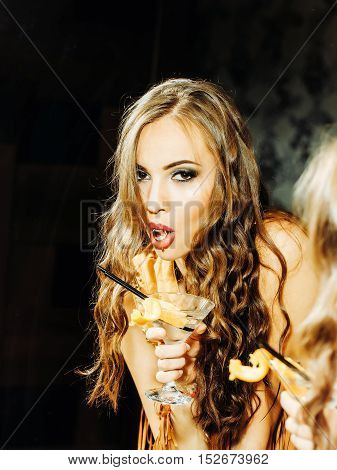Pretty girl cute party sexy fashion woman with long brunette curly hair with ice cube in mouth and cocktail looks in bathroom mirror