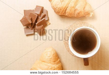 Two baked croissants with a cup of coffee and chocolate bars on white paper's surface.