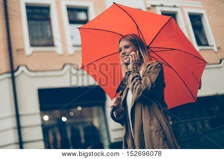 Good talk with friend. Low angle view of attractive young smiling woman carrying umbrella and talking on the mobile phone while walking down the street