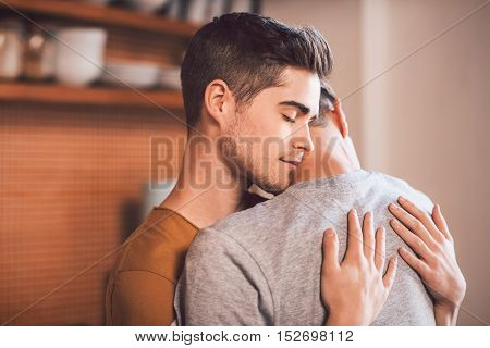 Affectionate young gay couple hugging each other with their eyes closed while standing in their kitchen in the morning