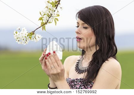 Sneezing hayfever girl is pained in green spring nature season. The beautiful young woman is pained by her allergy every year. She holds a tissue in her hands.