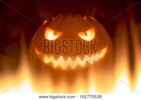 Bad carved halloween pumpkin in hot burning hell fire flames. The big helloween pumpkin has a mad face with glowing eyes and also a glow in its mouth and teeth. Perspective from bottom up.