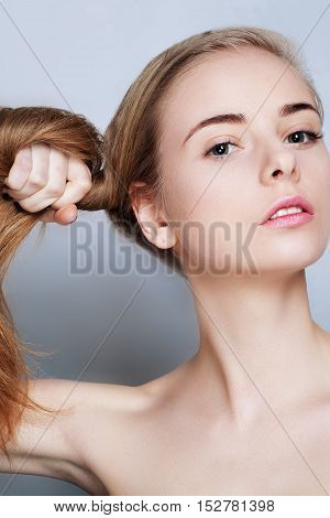 Beautiful blonde girl with long hair eyes thick eyebrows and naked shoulders holding hair at gray background beauty photo make up model portrait.