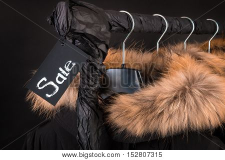 Lot Of Black Coats, Jacket With Fur On Hood Hanging  Clothes Rack.  Background. Sale Inscription.  F