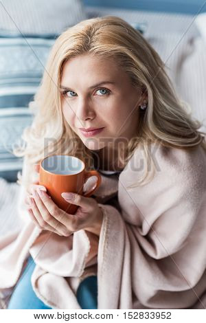 Creating warmth. Top view beautiful blond-haired woman covered with warm blanket holding big orange cup.