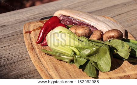 Natural raw duck breast with organic portabella mushrooms, red jalapeno pepper, green Chinese bok choy, green onion and ginger on a cutting board on a rustic wood farm table before going into duck pho soup.