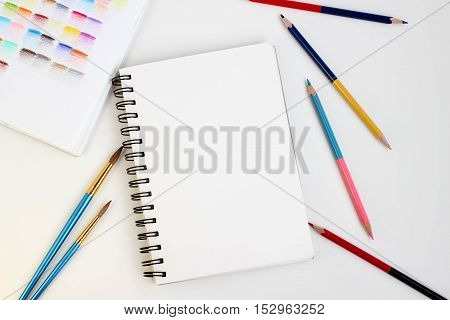 Top view of art supplies on white table. Colored pencils, paintbrushes and sketchbook.  May be used as product mockup.