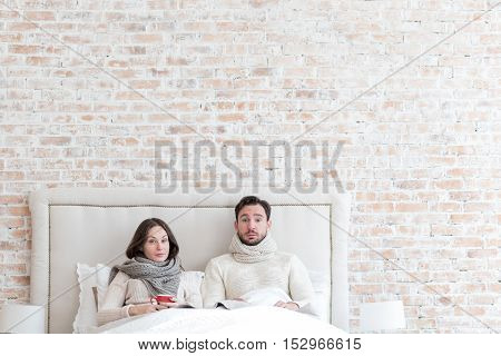 Being ill. Cheerless unhappy young couple lying together on a bed and looking in front of them while having cold