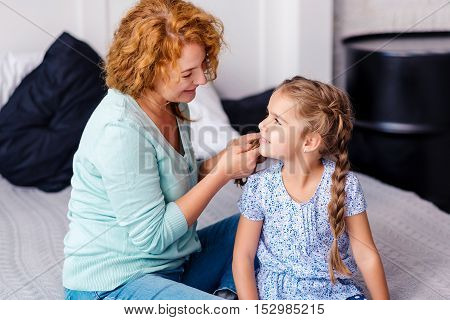 Making hairstyle. Cheerful smiling grandmother plaiting braids to her little granddaughter while having fun at home