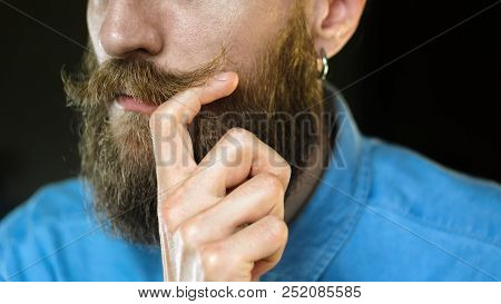 Bearded Man In A Blue Denim Shirt Twirling His Mustache With His Fingers. Portrait Of A Hipster On B