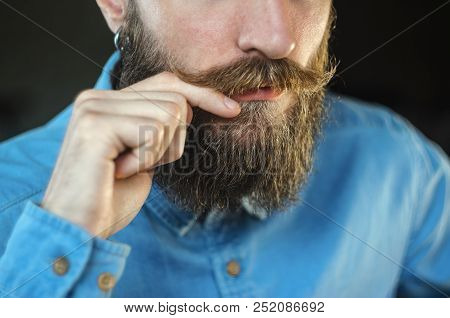Bearded Man In A Blue Denim Shirt Twirling His Mustache With His Fingers. Portrait Of A Hipster On B