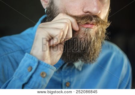 Bearded Man In A Blue Denim Shirt Twirling His Mustache With His Fingers. Portrait Of A Hipster On B