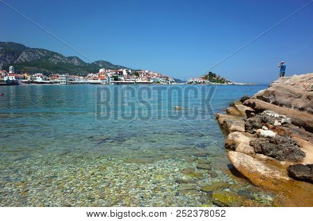 Kokkari, Samos island, Greece - 15 May, 2018: Man standing on rocky coast with stone bottom seen under crystal clean water with waterfront view of Kokkari fishing village