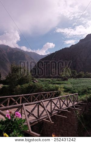 A Short Bridge Across The River In The Background Of High Mountains And Clouds