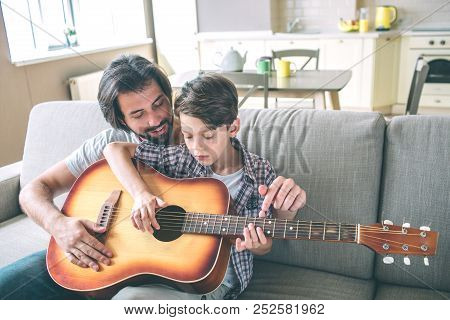 Boy Holds Big And Brown Guitar And Trying To Play. His Dad Is Sitting Behind Him And Help. They Are 