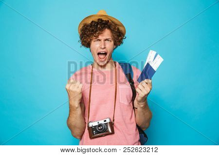 Image of photographer man 18-20 with curly hair wearing backpack and straw hat holding passport and tickets isolated over blue background
