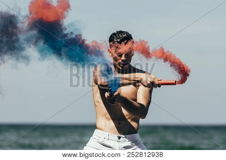Handsome Shirtless Man With Red And Blue Smoke Sticks In Front Of Ocean View