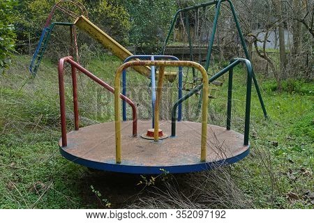 Playground In Overgrown Garden Of An Abandoned House. Rusty Merry-go-round Swings And Slide.