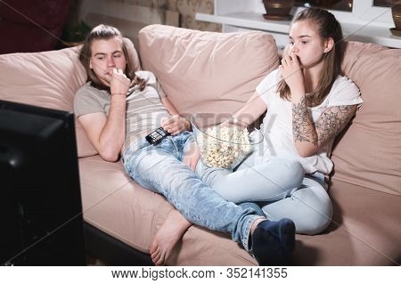Portrait Of A Long-haired Guy And Girl With Popcorn In Their Hands On The Couch Watching Tv. Family 