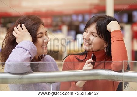 Indoor Shot Of Two Woman Talking Beside Clothes Rail In Shopping Mall, Look Happy, Females Gossiping