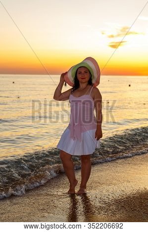 A Young Woman In A White Dress And Hat Stands On The Seashore At Sunset.
