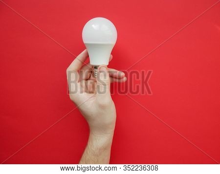 Female Hand Holds An Led Lamp On A Red Background. Energy Saving Concept, Alternative Energy Sources