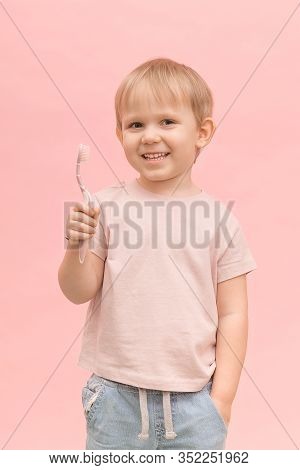 Happy Child Boy Blond European Appearance Holding A Toothbrush In His Hands On A Pink Background. Ve
