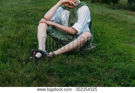 Boy In A White Shirt, Green Shorts And A Sweater Sits On Green Grass