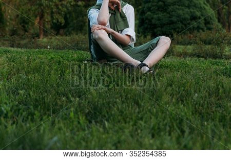 Boy In A White Shirt, Green Shorts And A Sweater Sits On Green Grass