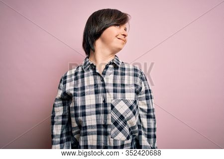 Young down syndrome woman wearing casual shirt over pink background looking away to side with smile on face, natural expression. Laughing confident.