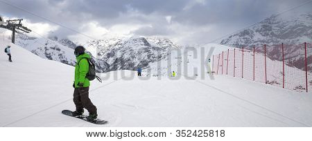 Snowy Ski Slope With Snowboarders And Skiers In High Mountains And Cloudy Sunlit Sky At Winter Gray 