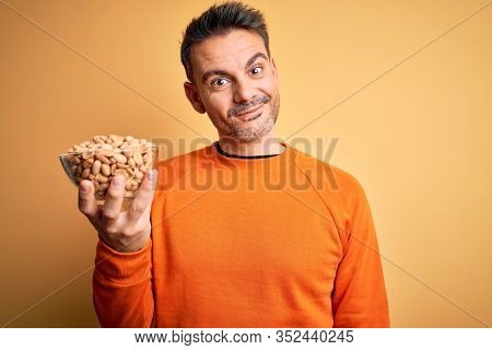 Young handsome man holding bowl with peanuts standing over isolated yellow background with a happy face standing and smiling with a confident smile showing teeth