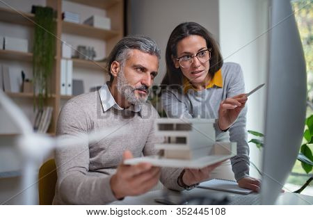 Architects With Model Of House Sitting At The Desk Indoors In Office, Working.