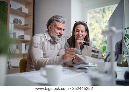 Architects With Model Of House Sitting At The Desk Indoors In Office, Working.