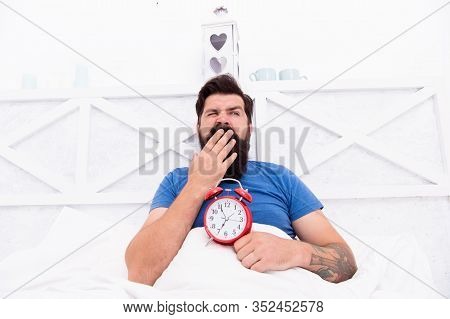 Stressed Man Yawning Takes Alarm Clock. Too Early. Sleepy Man Holding Alarm Clock In Bed. Bearded Ma