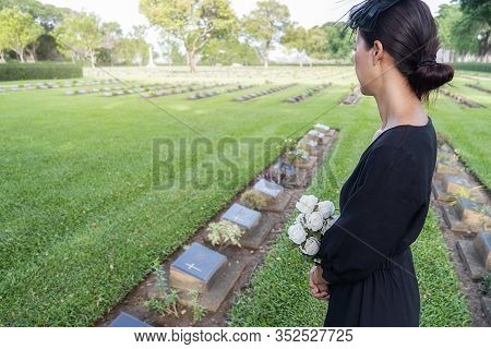 Mourning Young Woman Holding White Flowers At Her Family Grave In Beautiful Green Cemetery.