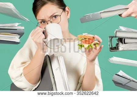Overworked Office Worker Drinks Coffee And Eats A Sandwich. At This Time, Hands With Documents And F