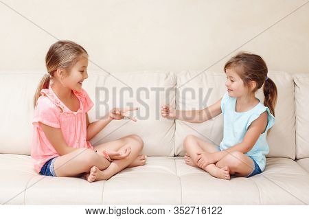 Two Girls Friends Playing Rock Paper Scissors Hand Game. Caucasian Children Sitting On Couch Playing