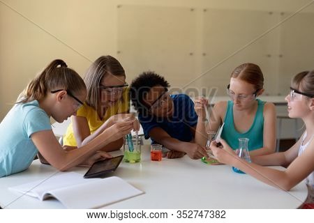 Front view of a multi-ethnic group of elementary school girls sitting at a table wearing safety glasses and using pipettes and beakers to do an experiment during a chemistry lesson, one girl holding a