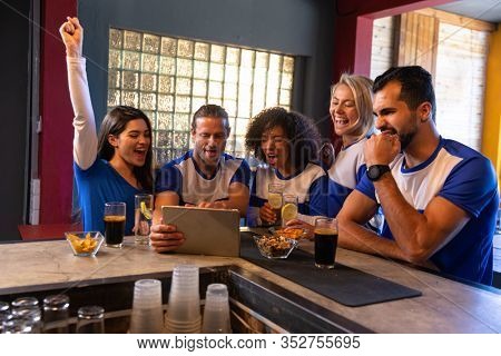 Front view of a group of five multi-ethnic male and female friends wearing team colours sitting at the bar in a pub, watching a sports match on a tablet computer and smiling, one woman wih her arm