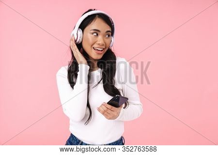Cheerful pretty young asian woman listening to music with wireless headphones isolated over pink background, holding mobile phone