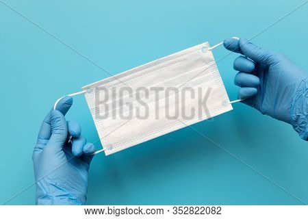 Hands Of Medic Wearing Blue Latex Gloves Holding Surgical Ear-loop Mask On Blue Background. Protecti