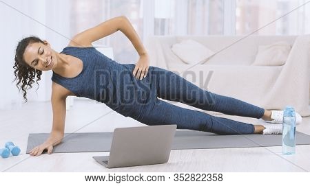 Happy Latin Woman Doing Side Plank Exercise While Watching Workout Video On Laptop At Home